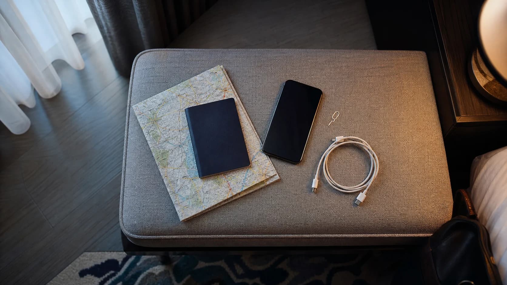 A traveler arranging a phone, passport cover, map, and charging cable on a hotel bench